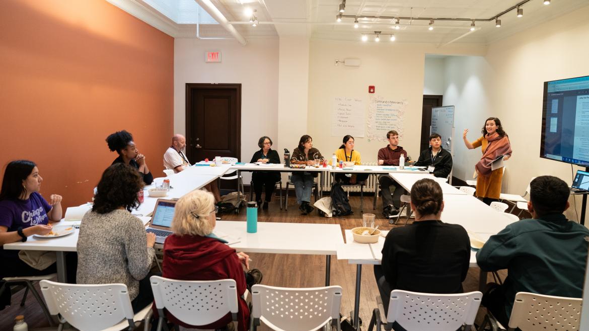 A group sit in a large square around tables while a presenter addresses them.